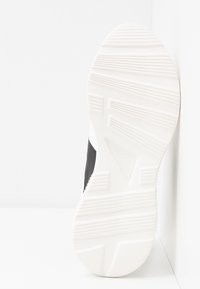 Zapatilla en blanco y negro que muestra una suela de goma texturizada con un patrón ondulado y características angulares para mayor tracción. Parte superior lisa en negro.