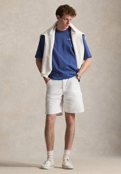 Μπλε βαμβακερό μπλουζάκι, λευκό σορτς και λευκά αθλητικά παπούτσια. Πουλόβερ περασμένο στους ώμους και ρολόι στον καρπό. Λεπτές υφές, χαλαρή εφαρμογή.