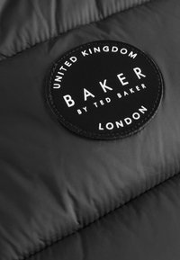 Crni platneni zakrpa s bijelim tekstom. Glasi: "Baker by Ted Baker, London, Ujedinjeno Kraljevstvo." Circularni dizajn s prošivenim rubom.
