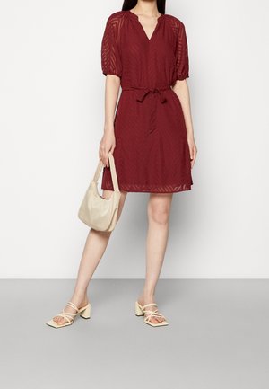 Femme portant une robe transparente bordeaux mi-longue à manches bouffantes, tenant un sac à main beige et chaussée de sandales à talons blocs beige avec brides.