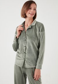 Camisa de pijama verde y aterciopelada con cuello, completo con botones en la parte delantera, mangas largas y botones negros. Combinada con pantalones de pijama a juego.