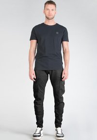 Camiseta negra de cuello redondo con un pequeño logo, combinada con pantalones cargo negros que tienen bolsillos laterales, y zapatillas blancas y negras.