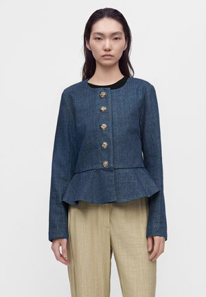 Modèle portant une veste en jean bleue ornée de boutons dorés et dotée d’un bas évasé, avec un pantalon beige ample, sur un fond uni.