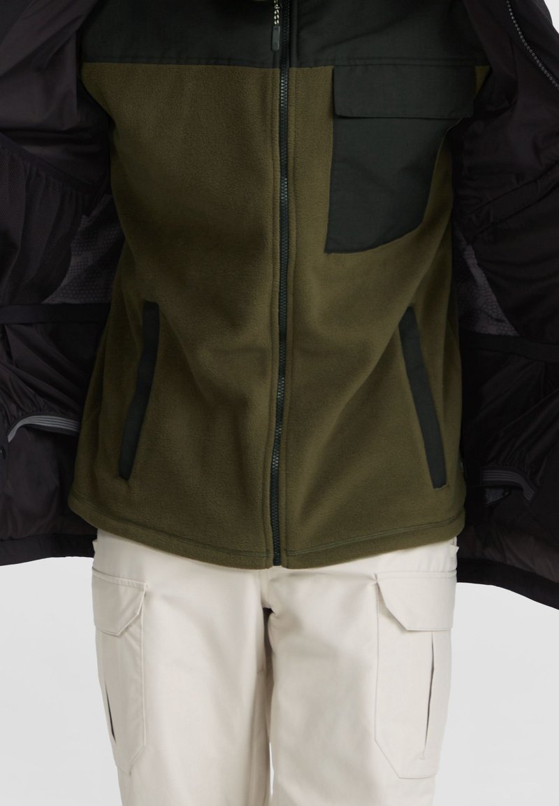 Haut du corps portant une veste polaire vert olive avec une poche poitrine noire et des poches latérales, associée à un pantalon cargo beige.