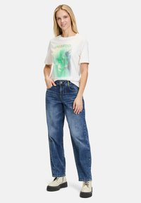 Weißes kurzärmeliges T-Shirt mit grünem Grafikdesign, kombiniert mit blauen Jeans. Das Model trägt cremefarbene Schnürstiefel mit einer dicken Sohle.
