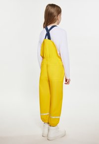 Geltonos neperšlampamos kombinezono kelnes su elastinga juosmens juosta, reguliuojamomis mėlynomis petnešomis ir atspindinčiais akcentais, kartu su baltomis aukštomis bateliais.