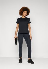 T-shirt atlético preto com logotipo nas mangas, combinado com leggings escuras e sapatilhas pretas. O modelo segura uma garrafa de água preta.
