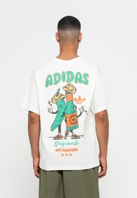 Hvid bomulds t-shirt med et farverigt grafisk motiv på ryggen, der viser tegneserie-jalapeño karakterer, orange detaljer og "ADIDAS" tekst.