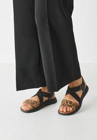 Crne i smeđe sandale s motivom leopard uzorka, teksturiranim trakama i glatkim đonom. Prekrižene podesive trake za sigurnu prilagodbu.
