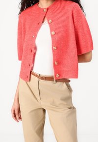 Koralový krátký cardigan s žebrovanou texturou, dřevěnými knofl íky a kulatým výstřihem, nošený přes bílý top, v kombinaci s béžovými kalhotami.