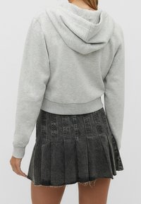Sudadera gris corta con bolsillo trasero y cordón, combinada con una falda negra con pliegues y dobladillo deshilachado. Material suave de mezcla de algodón.