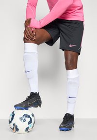 Αθλητική ένδυση Nike που περιλαμβάνει ένα ροζ μακρυμάνικο μπλουζάκι, μαύρα σορτς, λευκές κάλτσες με μπλε λεπτομέρειες και μαύρα ποδοσφαιρικά παπούτσια πάνω σε μια κανονική ποδοσφαιρική μπάλα.