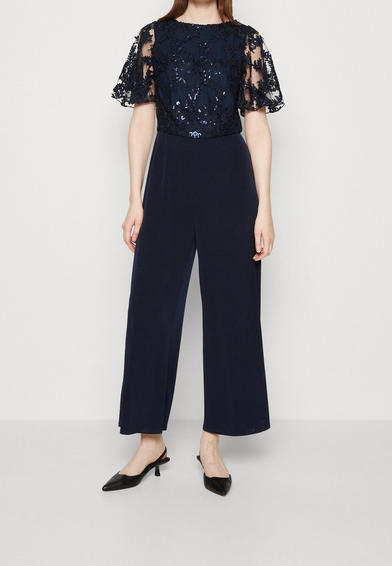 Femme portant une combinaison bleu marine avec des manches courtes en dentelle à sequins et un pantalon à jambes évasées, associée à des escarpins slingback noirs.