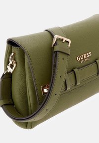 Borsa a tracolla in pelle verde oliva, con texture, hardware dorato, chiusura a fibbia e una lunga tracolla regolabile. Logo "GUESS" impresso.
