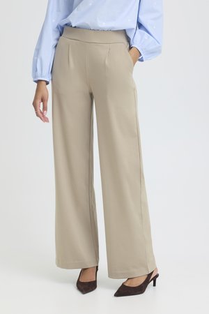 Nő bézs színű, bő szárú nadrágban, világoskék, fodros ujjú blúzban és sötétbarna, hegyes orrú magassarkú cipőben, kezét a zsebébe dugva.