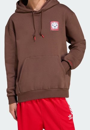 Person trägt einen braunen Hoodie mit einem Aufnäher eines Fußballballs und roten Kordelenden, kombiniert mit roten Adidas-Trainingshosen.