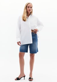 Camicia bianca con bottoni, pantaloni corte in denim blu con orlo sfrangiato, mule nere con tacco e un dettaglio decorativo. Un outfit semplice e casual.