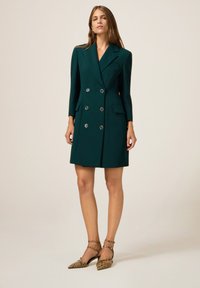 Frau trägt ein dunkelgrünes doppelt knöpfbares Blazer-Kleid mit silbernen Knöpfen und spitzen Pumps mit Schlangenleder-Muster und Knöchelriemen.
