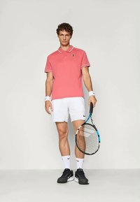 Rosa polo skjorte med hvite detaljer, kombinert med hvite shorts og svarte tennissko. Holder en tenniskølle, står foran en nøytral bakgrunn.