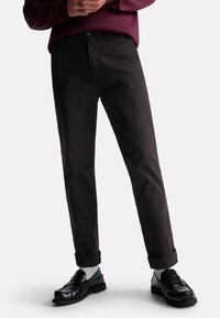 Chinos noirs en mélange de coton avec une coupe droite, dotés d'une fermeture à bouton, de poignets roulés, et associés à des mocassins noirs et des chaussettes blanches.