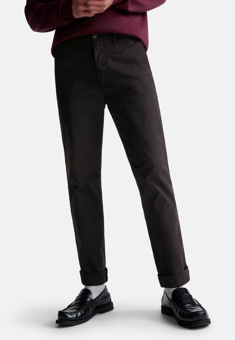Chinos noirs en mélange de coton avec une coupe droite, dotés d'une fermeture à bouton, de poignets roulés, et associés à des mocassins noirs et des chaussettes blanches.