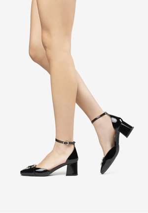 Zapatos negros de charol con tacón grueso, correa en el tobillo y hebilla decorativa en el empeine. Acabado brillante, diseño de punta afilada.