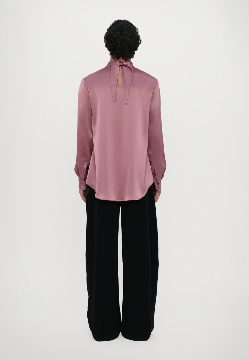 Blouse en satin rose avec manches longues, détail de lien à l'arrière, encolure à empiècement, associée à un pantalon noir ample, texture lisse, coupe décontractée.