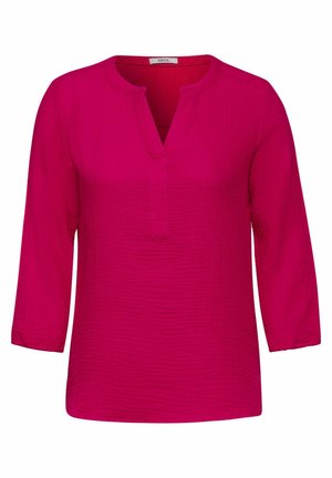 Blouse rose vif à manches longues avec un col rond et une fente à l'avant, tissu légèrement texturé et petite étiquette au col.