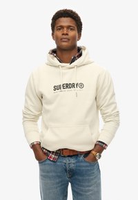 Felpa con cappuccio beige con logo "SUPERDRY", dotata di tasca frontale, indossata sopra una camicia a scacchi, abbinata a jeans blu e un orologio marrone.