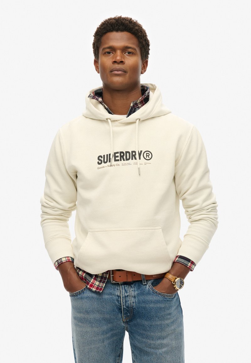 Felpa con cappuccio beige con logo "SUPERDRY", dotata di tasca frontale, indossata sopra una camicia a scacchi, abbinata a jeans blu e un orologio marrone.