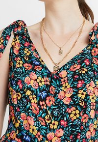 Robe à motifs floraux sur fond noir, avec des fleurs colorées en orange, jaune et bleu ; bretelles nouées et décolleté en V profond. Colliers en or superposés visibles.
