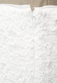 Κοντινή λήψη λευκής δαντέλας με μερικώς φερμουάρ στη μέση, που έχει ένα μικρό λευκό τσιμπιδάκι, σε μπεζ φόντο.