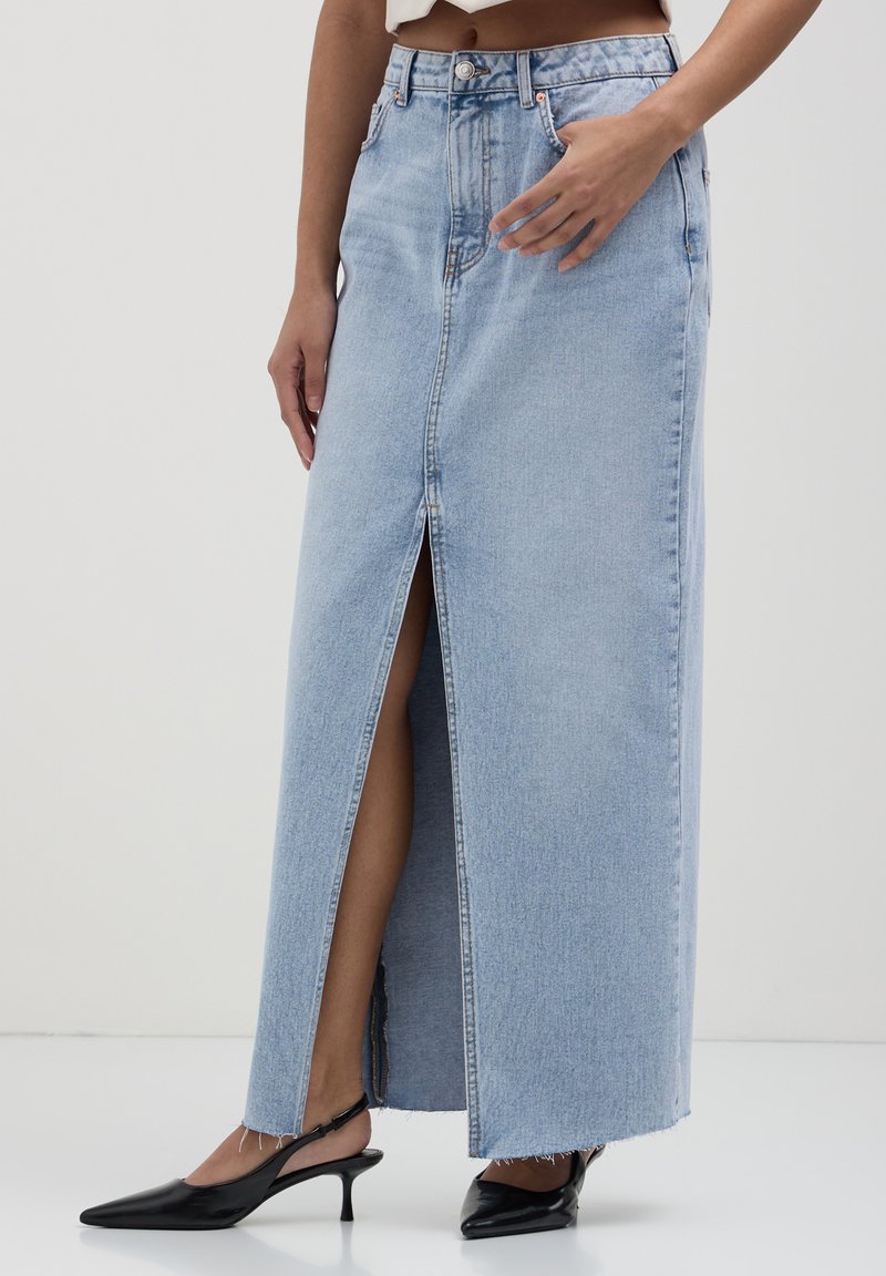 Jupe en denim bleu clair avec une fente avant, taille haute et ourlet brut. Assortie à des talons slingback noirs à bout pointu.