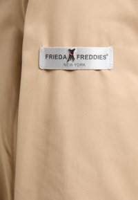Beżowy materiał o gładkiej fakturze z naszytą metką. Metka zawiera napis "FRIEDA FREDDIES" w czarnym kolorze oraz "NEW YORK" poniżej.