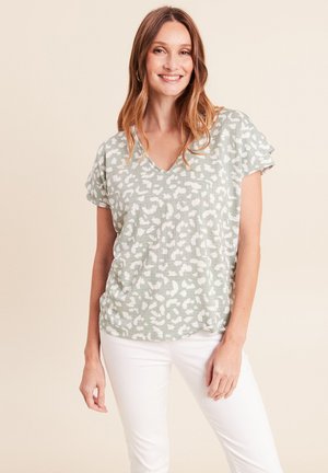Lachende vrouw met schouderlang haar, gekleed in een groen-wit patroon V-hals top en witte broek, staand tegen een beige achtergrond.