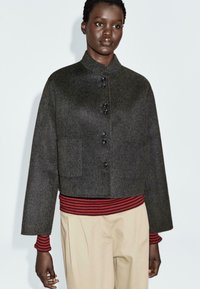 Giacca di lana grigia scura con bottoni sopra una maglietta a righe rossa e nera, abbinata a pantaloni beige, indossata da una persona in piedi su uno sfondo semplice.