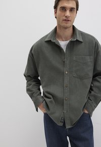 Olivgrünes Cordhemd mit Knöpfen, langen Ärmeln, Brusttasche und Holzknöpfen. Über einem weißen T-Shirt getragen und mit blauen Jeans kombiniert.