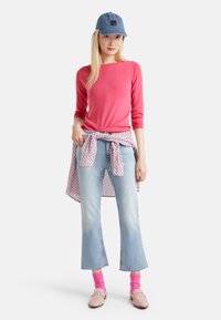 Maglione rosa a maniche lunghe, jeans a campana blu chiaro, calzini rosa, mocassini rosa chiaro e un cappellino blu con un patch logo. Camicia a motivo nodi annodata in vita.