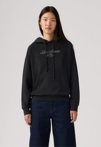 Sudadera negra con bolsillo frontal, capucha con cordón y logo bordado. Combinada con vaqueros azul de cintura alta. Textura de tejido suave.