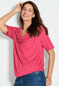 Top a maniche corte di un brillante colore rosa, con scollatura drappeggiata e tessuto leggero, abbinato a pantaloni in denim. Texture liscia e vestibilità casual.