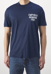 Námořnická modrá bavlněná tričko s krátkými rukávy, s bílým vyšitým textem "EMPORIO ARMANI 1981" na oblasti levého prsou.