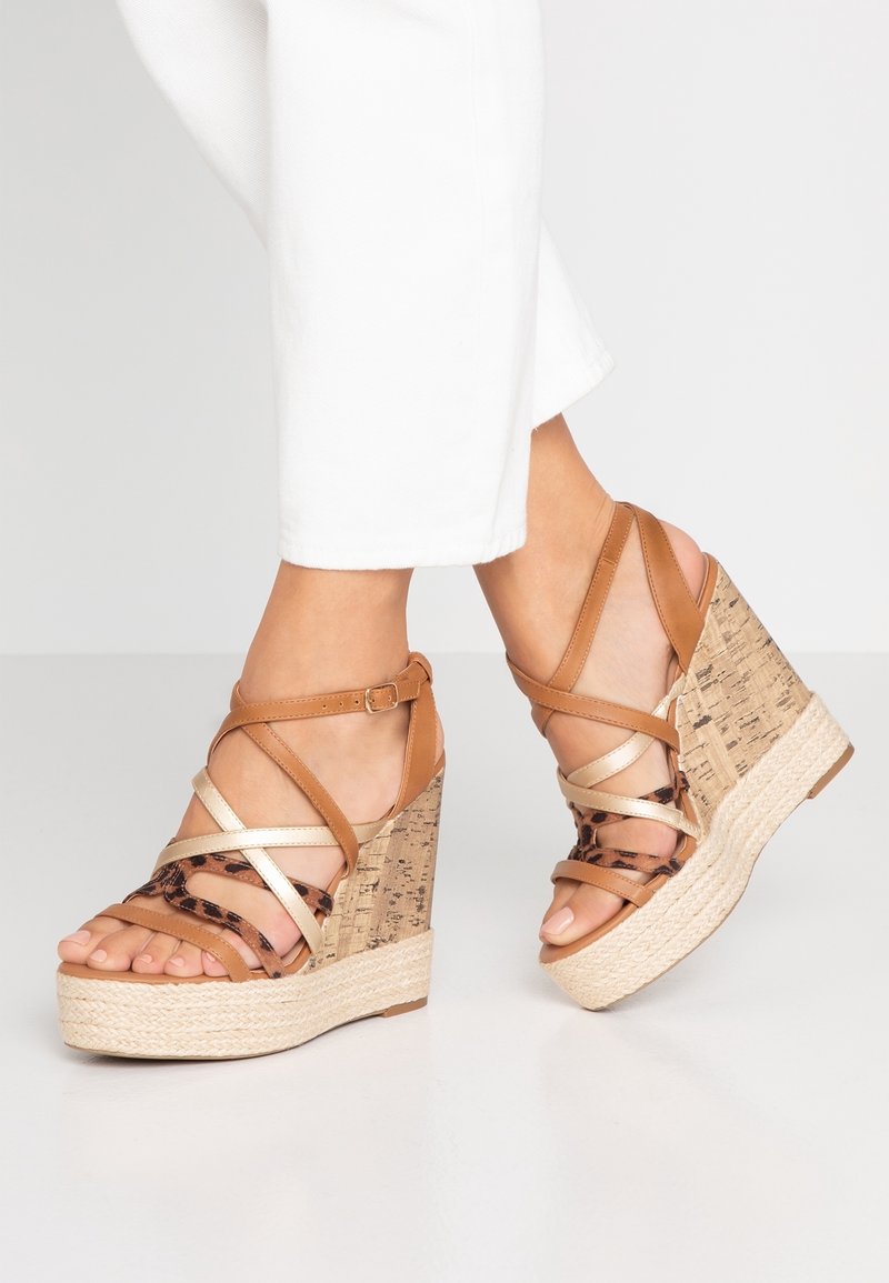 Jambes vêtues d'un pantalon blanc et de sandales compensées couleur fauve avec semelle en liège, design multi-lanières incluant des accents léopard et métalliques, sur un sol blanc.