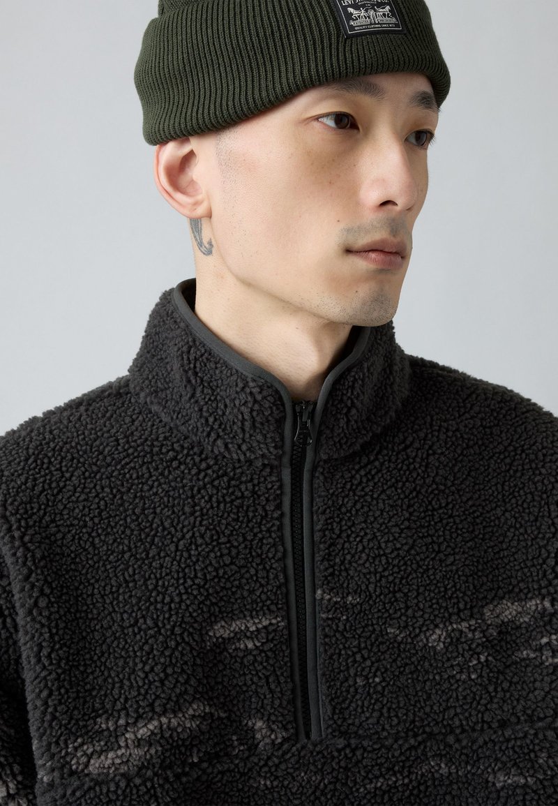 Mann trägt eine grüne Strickmütze und eine schwarze, strukturierte Fleecejacke mit Reißverschluss, schaut zur Seite mit einem sichtbaren Nackentattoo.