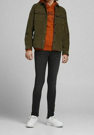 Giacca di velluto verde oliva indossata sopra una felpa con cappuccio arancione, abbinata a jeans skinny neri e sneakers bianche. Tasche frontali di rilievo sulla giacca.