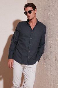 Camisa de botones de color gris oscuro con cuello, mangas largas y botones blancos, combinada con pantalones de tono claro frente a una pared texturizada.