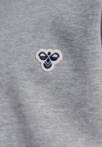 Grau gestrickter Stoff mit einem kleinen bestickten Bienenlogo in Marineblau und Weiß, platziert auf der oberen linken Seite.