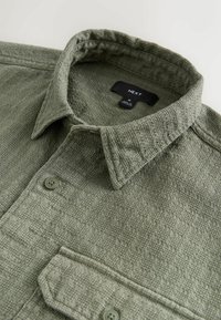 Oliwkowo-zielona koszula na guziki z teksturowanej tkaniny, z kołnierzykiem i przednią kieszenią, wyposażona w dwa zielone guziki na patce do zamknięcia.