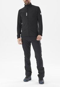 Homme portant une veste polaire zippée noire et un pantalon de randonnée noir avec des poches zippées, se tenant contre un fond blanc.