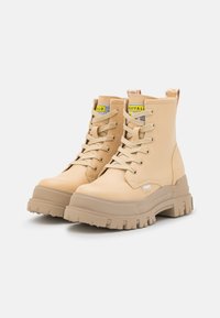 Beżowe botki sięgające kostki, wykonane z gładkiego materiału syntetycznego, z przodu sznurowane, z pętlą do zakładania i grubą teksturowaną podeszwą.