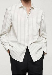 Chemise blanche à manches longues avec boutons et fines rayures verticales grises, portée non rentrée, mains dans les poches, associée à un pantalon noir.